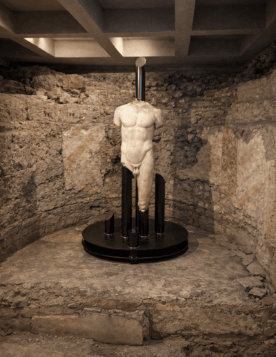 Statua dell’Atleta esposta su base circolare con supporto strutturale in ambiente archeologico restaurato