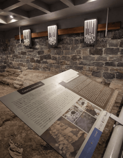 Allestimento museale del Portico dell’Atleta a Catania con pannello didattico inclinato su area archeologica e illuminazione integrata su parete in pietra lavica