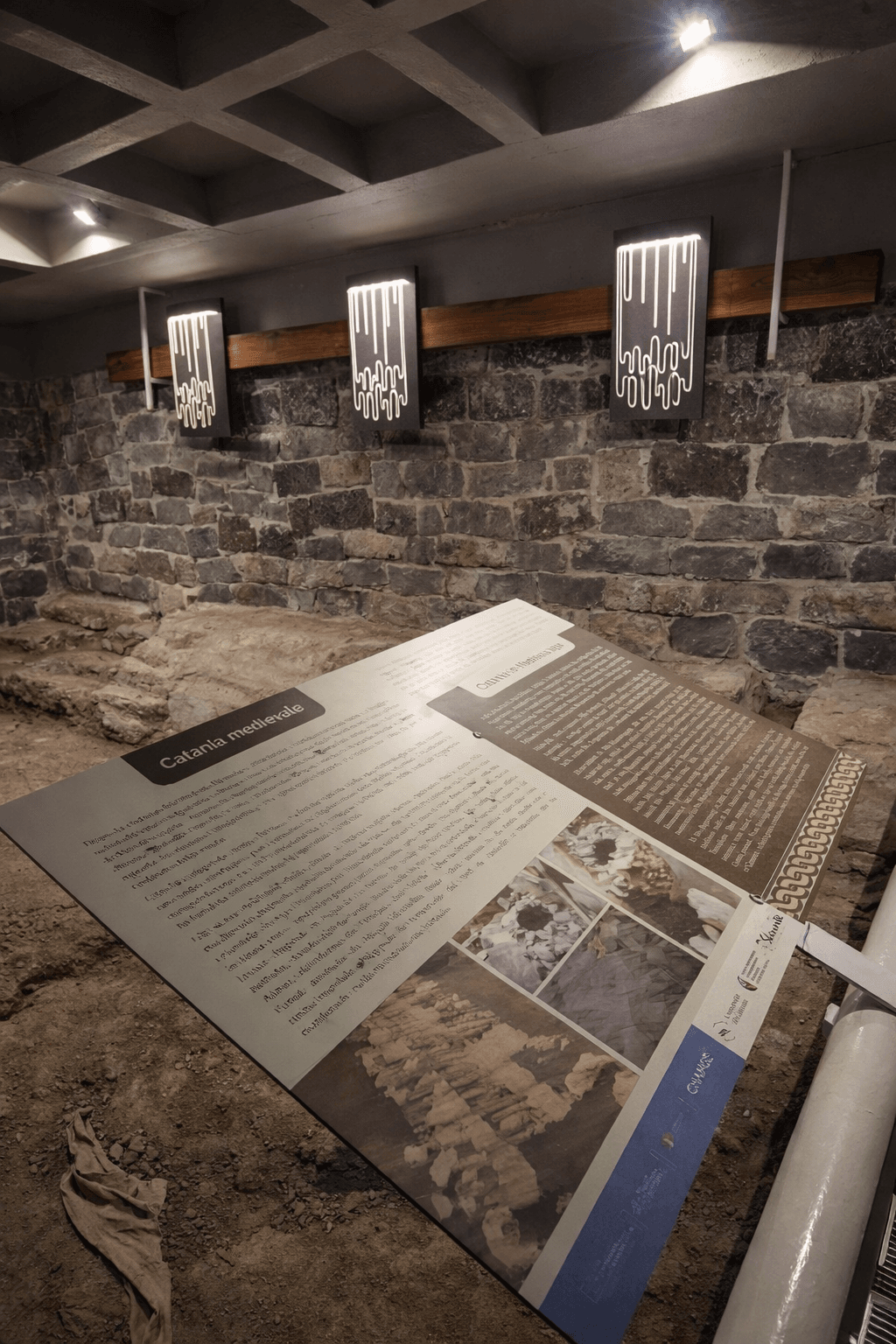 Allestimento museale del Portico dell’Atleta a Catania con pannello didattico inclinato su area archeologica e illuminazione integrata su parete in pietra lavica