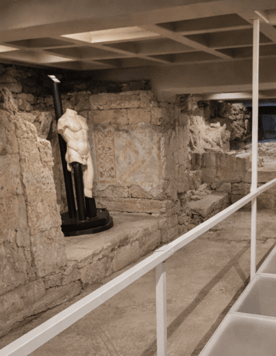 Passerella contemporanea in struttura metallica bianca all’interno del Portico dell’Atleta con resti archeologici e statua antica su basamento moderno.