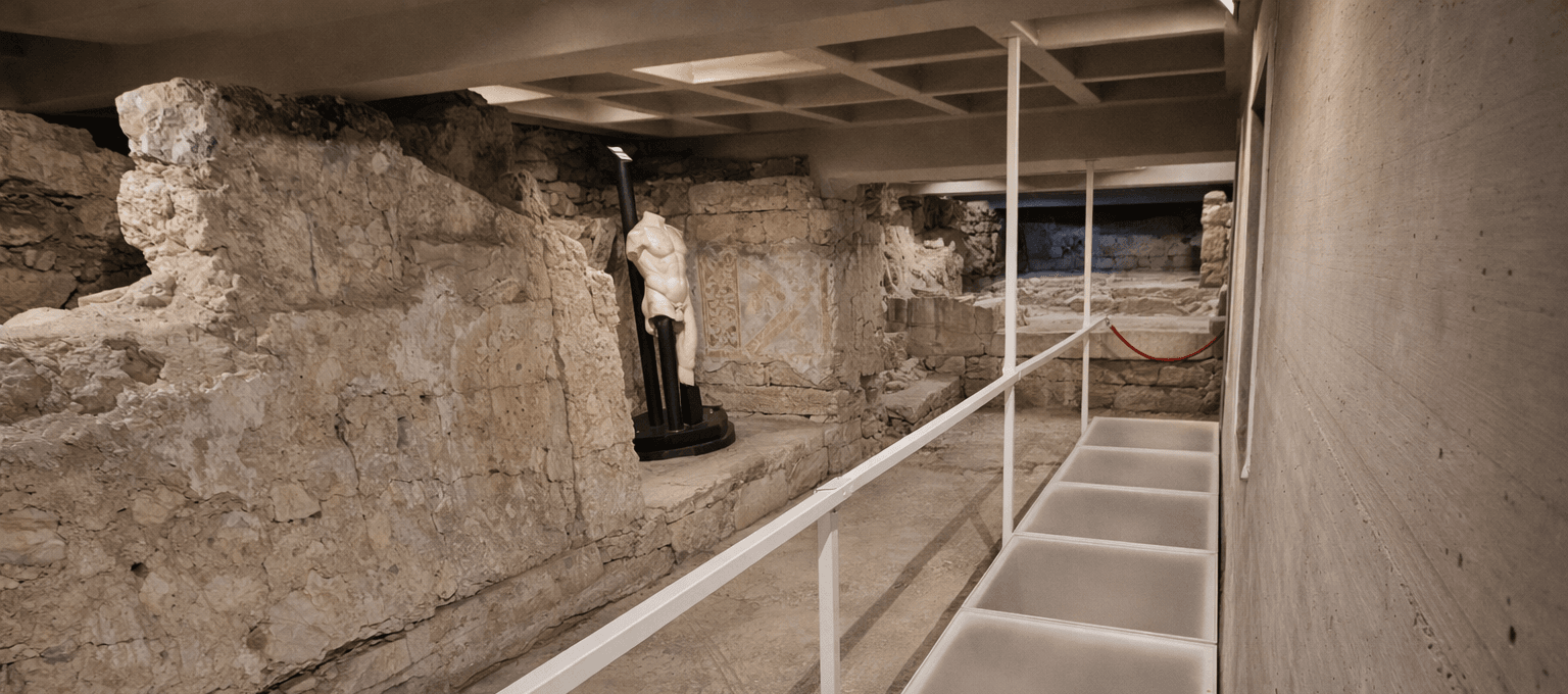 Passerella contemporanea in struttura metallica bianca all’interno del Portico dell’Atleta con resti archeologici e statua antica su basamento moderno.